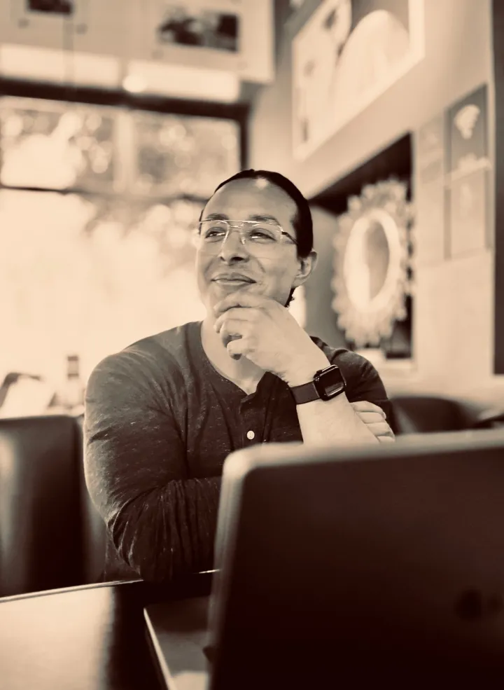 René Velarde smiling, chin in hand, at a table with a laptop