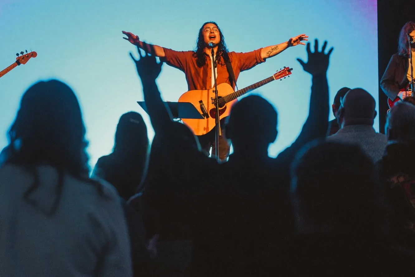 René Velarde leading worship on stage with an acoustic guitar, arms open wide, as people in the room raise their hands